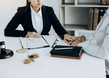 Two people in formal attire sit across from each other at a desk with legal documents, a gavel, and scales of justice, likely consulting with their workers compensation lawyer. They hold hands in a gesture of support. Books are visible in the background, suggesting a legal or professional setting. | FintechZoom