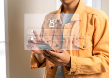 A person in a yellow jacket uses a smartphone, engaged in digital onboarding. A virtual login interface with user and lock icons is overlayed in front of them, symbolizing security and connectivity. | FintechZoom