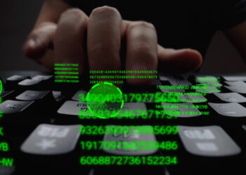 A close-up of a hand typing on a keyboard, with green digital numbers and codes overlaid, resembling a computer hacking scene. The dark background emphasizes the glowing green text, hinting at underlying security strategies in action. | FintechZoom