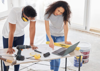 A man and woman collaborate on a home renovation project, focusing on home improvements. They examine a laptop and point to a blueprint on the table, surrounded by tools like a drill, hammer, level, and tape, all essential for bringing their vision to life. | FintechZoom