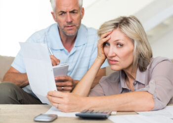 A concerned couple reviews documents at a table, the woman glancing worriedly at the camera while holding a paper. The man writes in a notepad, possibly noting down details from debt collectors. A calculator and smartphone are on the table, underscoring their financial stress. | FintechZoom