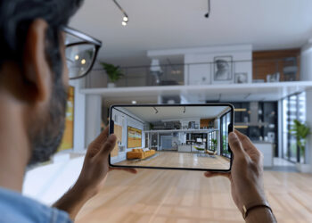 A person holding a tablet in a modern living room uses AI-powered real estate solutions to capture the same room through the screen. Featuring wooden flooring, a couch, large windows, and a loft area, the space is adorned with various plants and framed artworks. | FintechZoom
