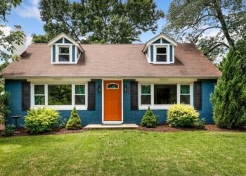 A charming blue house with a brown roof and two dormer windows. The front features an orange door, flanked by symmetrical white-framed windows, and surrounded by well-maintained green shrubs and grass. Tall trees are visible in the background. | FintechZoom