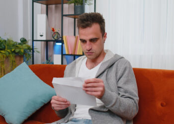 A man in a gray hoodie and white t-shirt sits on an orange sofa, intently reviewing a denial for his workers' compensation claim. Behind him, there's a bookshelf with plants and colorful items, enhancing the cozy ambiance of the living room. | FintechZoom