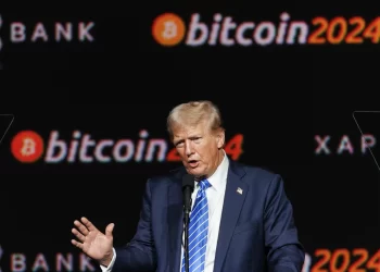Donald Trump, speaks into a microphone. Behind him, the words "Bitcoin 2024" are prominently displayed, surrounded by logos and other text on a dark background. | FintechZoom