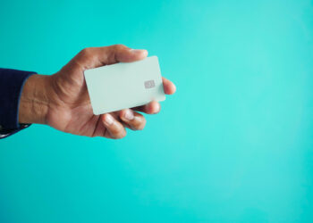 A person holds a blank white credit card with a visible chip against a bright turquoise background. | FintechZoom