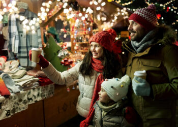 A family of three, bundled in winter clothes, delight in a festive Christmas market. They hold hot drinks and smile while admiring holiday decorations and lights. In the background, a stall displays colorful scarves and mittens. | FintechZoom
