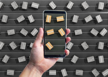 A hand holds a smartphone, enhanced with mobile proxies, displaying a close-up of a conveyor belt with small cardboard boxes. The phone screen shows a magnified view, while the background features numerous similar boxes on a larger conveyor belt. | FintechZoom