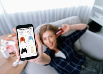A woman relaxes on a couch, smartphone in hand, displaying an online shopping app with various clothing items and tips on how to know your size. Smiling as she holds a credit card, she's surrounded by a bright, modern living space. | FintechZoom