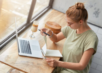 A woman with glasses is sitting by a window at a wooden table, working on her blog from a laptop. She's jotting notes in a notebook with a pen beside her glass of iced coffee. Dressed in a green shirt, her hair is neatly tied up in a bun. | FintechZoom