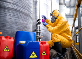 In a scene highlighting industrial chemical safety, a person in a yellow hazmat suit and gas mask kneels in an industrial setting, examining a small vial of purple liquid. Red and blue biohazard-labeled containers are scattered in the foreground, while large metallic tanks loom in the background. | FintechZoom