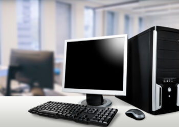 A low-end PC setup sits on a white desk in a modern office environment, complete with a monitor, keyboard, and tower. Blurred office furniture provides the backdrop, emphasizing a streamlined yet accessible workspace. | FintechZoom