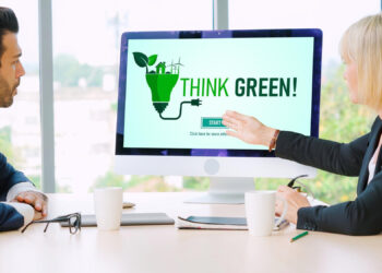 Two professionals sit at a desk in an office, discussing a computer screen displaying the words "THINK GREEN!" with eco-friendly imagery, including plants and a green light bulb. Coffee cups and documents are on the table. | FintechZoom