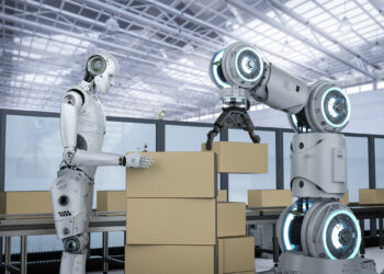 A humanoid robot collaborates with a robotic arm to stack cardboard boxes in a warehouse setting, exemplifying how collaborative robots are redefining automation in manufacturing. The environment features a high ceiling with large windows, allowing natural light to illuminate the scene. | FintechZoom