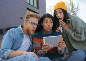 Three people, embodying the Gen Z vibe, sit outdoors, eyes wide with surprise as they huddle around a tablet. One sports glasses and a denim shirt, another a colorful sweater, and the third rocks a yellow beanie with a green jacket. Everywhere you look, their intrigue is unmistakable. | FintechZoom