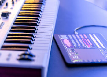 A close-up image of a music production setup featuring a keyboard synthesizer and tablet using LMS Music Software. The tablet displays the app with waveforms and controls, connected to the synthesizer, all illuminated in a cool blue tone. | FintechZoom