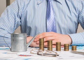 A person in a blue striped shirt and tie sits at a table with folded hands. In front are stacks of coins increasing in height, a small watering can, glasses, and various financial charts and graphs—an ideal scene for someone investing in sector-specific mutual funds. | FintechZoom