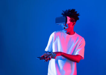 A person wearing virtual reality goggles smiles and points at a smartphone in their hand. The scene is illuminated with blue and pink lighting, creating a futuristic atmosphere. They are dressed in a plain white T-shirt. | FintechZoom