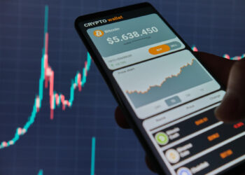 A person holds a smartphone displaying a cryptocurrency wallet app, showcasing Bitcoin at $5,638.450. The screen features a price chart and buy/sell options, typical of crypto trading platforms. In the background, a larger financial chart is visible on a computer monitor. | FintechZoom