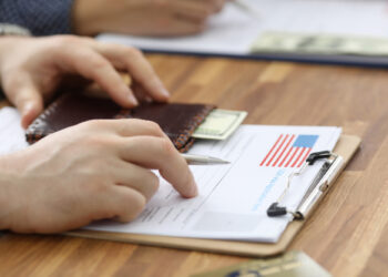 A person is filling out a form on a clipboard adorned with an American flag design, possibly related to US unemployment rises. A hand grips a pen while a wallet nearby reveals a partially visible dollar bill, and another individual works in the background. | FintechZoom