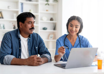 A healthcare professional in scrubs sits with a man in casual attire at a desk, smiling and explaining health insurance details on a laptop. The man listens attentively amid bookshelves in the background. | FintechZoom