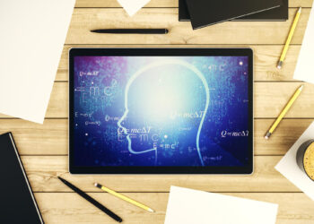 A tablet on a wooden desk displays scientific equations against a blue background with an outline of a human head, reminiscent of IQ testing. Sheets of paper, pencils, a pen, a notebook, and a steaming cup of coffee surround the scene. | FintechZoom