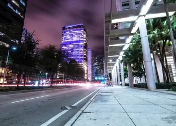 City street at night with skyscrapers lit in purple and white lights. Blurred car lights create streaks on the road, reminiscent of bustling Miami nights. Palm trees line the sidewalk, and the sky is a dark purple with clouds. The scene is vibrant and dynamic, echoing the city's lively pulse.
. | FintechZoom