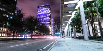 City street at night with skyscrapers lit in purple and white lights. Blurred car lights create streaks on the road, reminiscent of bustling Miami nights. Palm trees line the sidewalk, and the sky is a dark purple with clouds. The scene is vibrant and dynamic, echoing the city's lively pulse.
. | FintechZoom