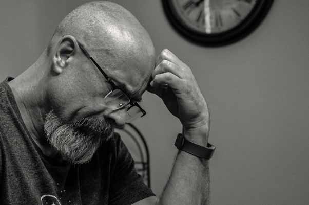 A black and white photo of a man with glasses and a beard, seated with his hand on his forehead in a thoughtful pose. A wall clock is visible in the background. | FintechZoom