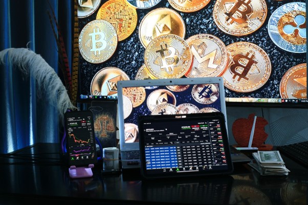 A desk setup featuring a laptop, tablet, and smartphone displaying cryptocurrency trading charts. The background shows a large screen with images of various cryptocurrency coins, including Bitcoin and Monero. A stack of cash is visible on the right. | FintechZoom