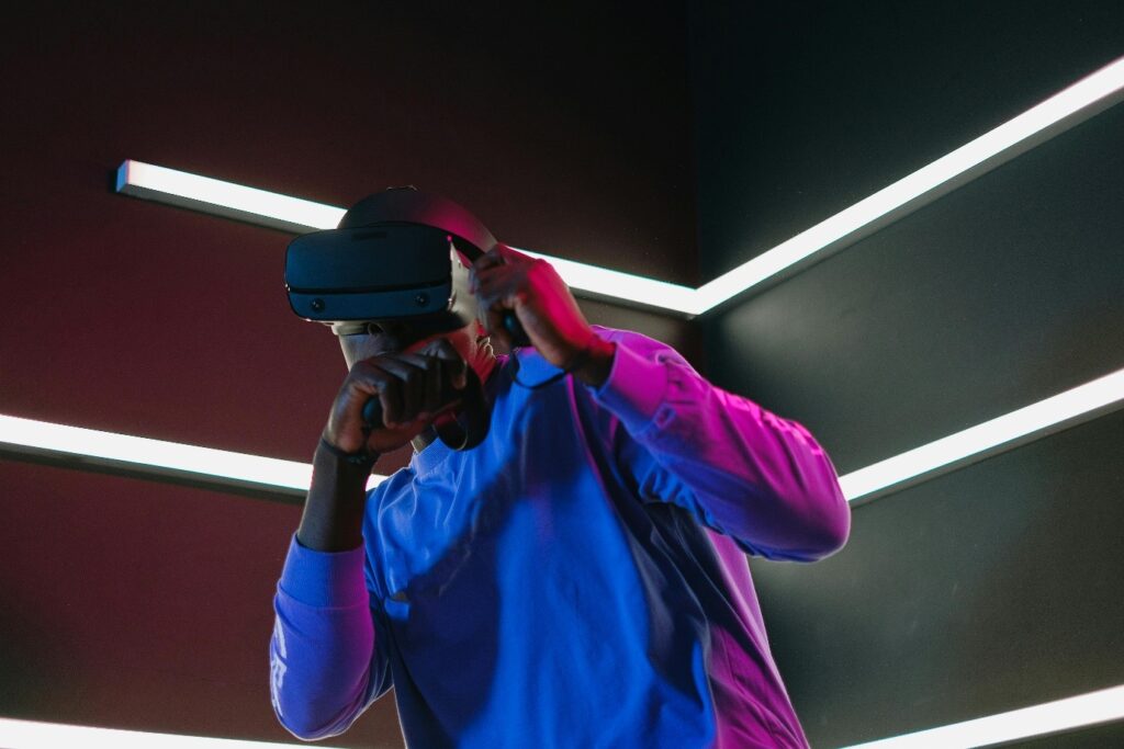 A person wearing a virtual reality headset stands in a dark room with vibrant neon lights on the walls. The individual appears engaged with the VR experience, dressed in a long-sleeved blue shirt. | FintechZoom