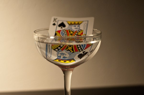 A king of spades playing card rests in a clear, shallow champagne glass, reminiscent of slot machines where luck is both submerged and reflected. Its partial immersion creates an intriguing reflection against a plain background, echoing the allure of chance. | FintechZoom