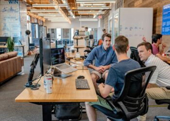 A group of three people sit and discuss around a desk in a modern, open office space, brainstorming ideas to develop a mobile application. One person types on a laptop, while the others listen attentively. The office boasts a wooden interior design with bright lighting and several desks in the background. | FintechZoom