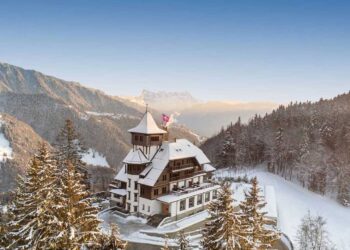 A grand alpine lodge, resembling a luxury rehab retreat, is surrounded by snow-covered trees with majestic mountains in the background under a clear blue sky. This scene captures a serene winter landscape. | FintechZoom