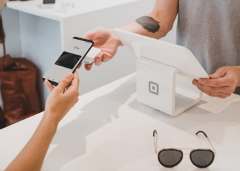 A person holds a smartphone displaying a payment app near a white point-of-sale device. Another person operates the device. Sunglasses and a brown bag are visible on a white surface. | FintechZoom