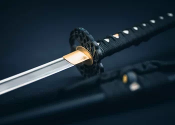 Close-up of a Samurai sword, showcasing its sharp, polished blade and intricately detailed handle against a dark background. A black scabbard is partially visible beside this elegant katana. | FintechZoom