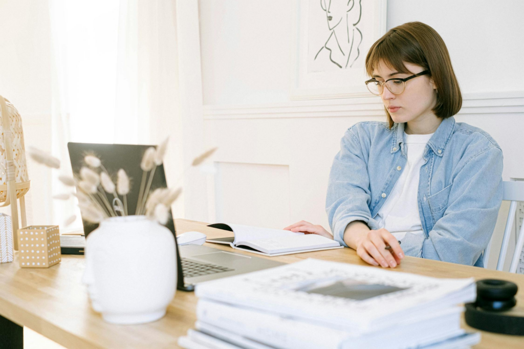 A person wearing glasses and a denim shirt is sitting at a desk reading a book. A laptop, a vase with pampas grass, and several books are on the table. The background features a framed line drawing on the wall. | FintechZoom