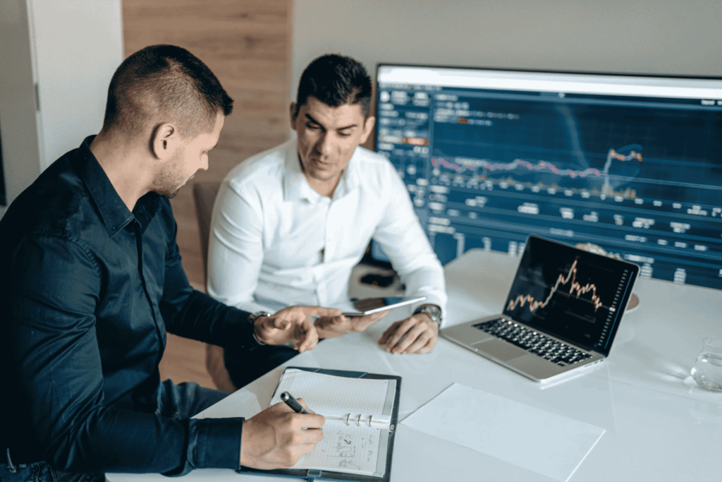 Two men in an office discussing financial charts. One is writing in a notebook, and the other is holding a tablet. A large monitor and a laptop display graphs and data. They are seated at a white table, focused on their work. | FintechZoom