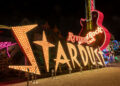 Neon signs in a nighttime scene featuring large, illuminated letters spelling "Stardust," with a giant guitar and the words "Hard Rock Cafe" in the background. Other neon lights and signs are partially visible. | FintechZoom