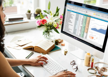 A woman works at a desk with accounts receivables management software open on her computer, displaying spreadsheets and charts. The desk features a vase of flowers, an open book, glasses, and personal items. Natural light streams through a window in the background. | FintechZoom