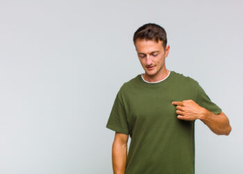 Man in a green t-shirt points to his chest against a light gray background. With short brown hair and a neutral expression, he stands with his right hand raised, emphasizing the t-shirt as a statement piece. | FintechZoom