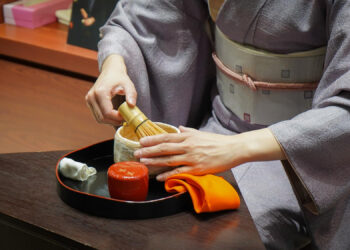 A person in a traditional Japanese kimono is preparing matcha tea, embracing elements reminiscent of traditional Korean healing practices. They use a bamboo whisk to mix the tea in a bowl on a tray, where an orange cloth and small container also rest. | FintechZoom