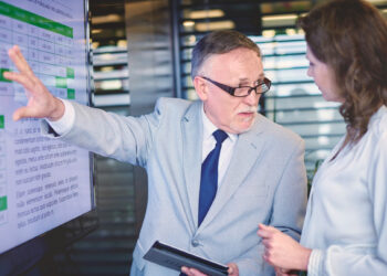 An older man in a gray suit and glasses explains information on a screen to a woman with long brown hair in a white blouse. The screen displays a spreadsheet. The man holds a tablet, gesturing toward the screen. | FintechZoom