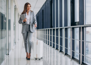 A woman in a gray suit strides down a bright corridor, epitomizing business travel. She pulls a white suitcase and holds a laptop, her black heels clicking confidently. Smiling, large windows on her left let in natural light, illuminating her journey. | FintechZoom