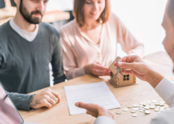 A couple sits at a table completing a real estate transaction, guided by a mortgage broker. They receive keys over a tiny house model and contract. Coins are spread on the table, while a laptop hums quietly in the corner. | FintechZoom