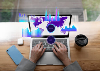 A person types on a laptop showcasing data visualizations, including graphs and charts, overlaid on a world map. Nearby, a coffee cup, phone, and AirPods case rest on a wooden desk—a scene reminiscent of the emerging future of real-time digital transactions. | FintechZoom