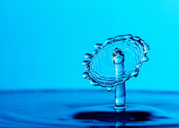 A close-up of a water droplet collision creating an umbrella-like splash against a blue background. The surface tension forms intricate shapes as the droplet rises and spreads out, capturing a moment of fluid dynamics. | FintechZoom