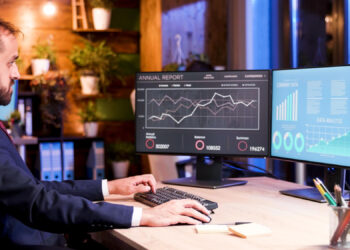 A man in a suit works at a desk with two computer monitors, utilizing financial management software to analyze charts and graphs related to annual financial reports. The office boasts a modern design, complemented by lush plants and sleek shelves in the background. | FintechZoom