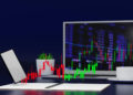 A laptop displaying financial charts with red and green candlesticks is placed on a desk. A tablet, a notebook, a potted plant, and a coffee cup on a saucer are also on the desk, set against a dark blue background. | FintechZoom