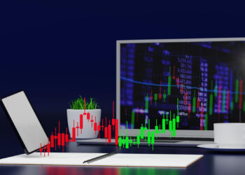 A laptop displaying financial charts with red and green candlesticks is placed on a desk. A tablet, a notebook, a potted plant, and a coffee cup on a saucer are also on the desk, set against a dark blue background. | FintechZoom